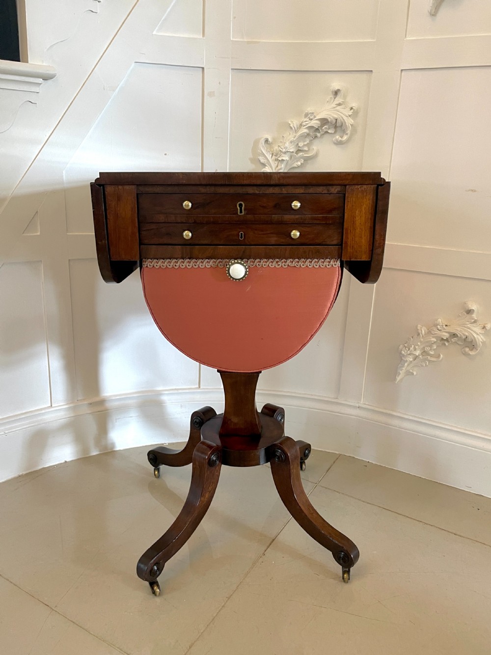An antique Regency rosewood sewing table featuring small drop leaves, drawers with brass handles, a terracotta fabric work basket below, and a pedestal base with four splayed legs on castors. View XX