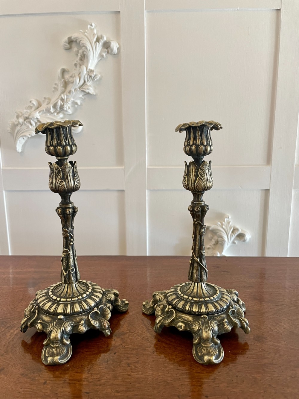 A pair of ornate brass candlesticks featuring intricate foliage carving, fluted stems, and scalloped candle sockets. View X