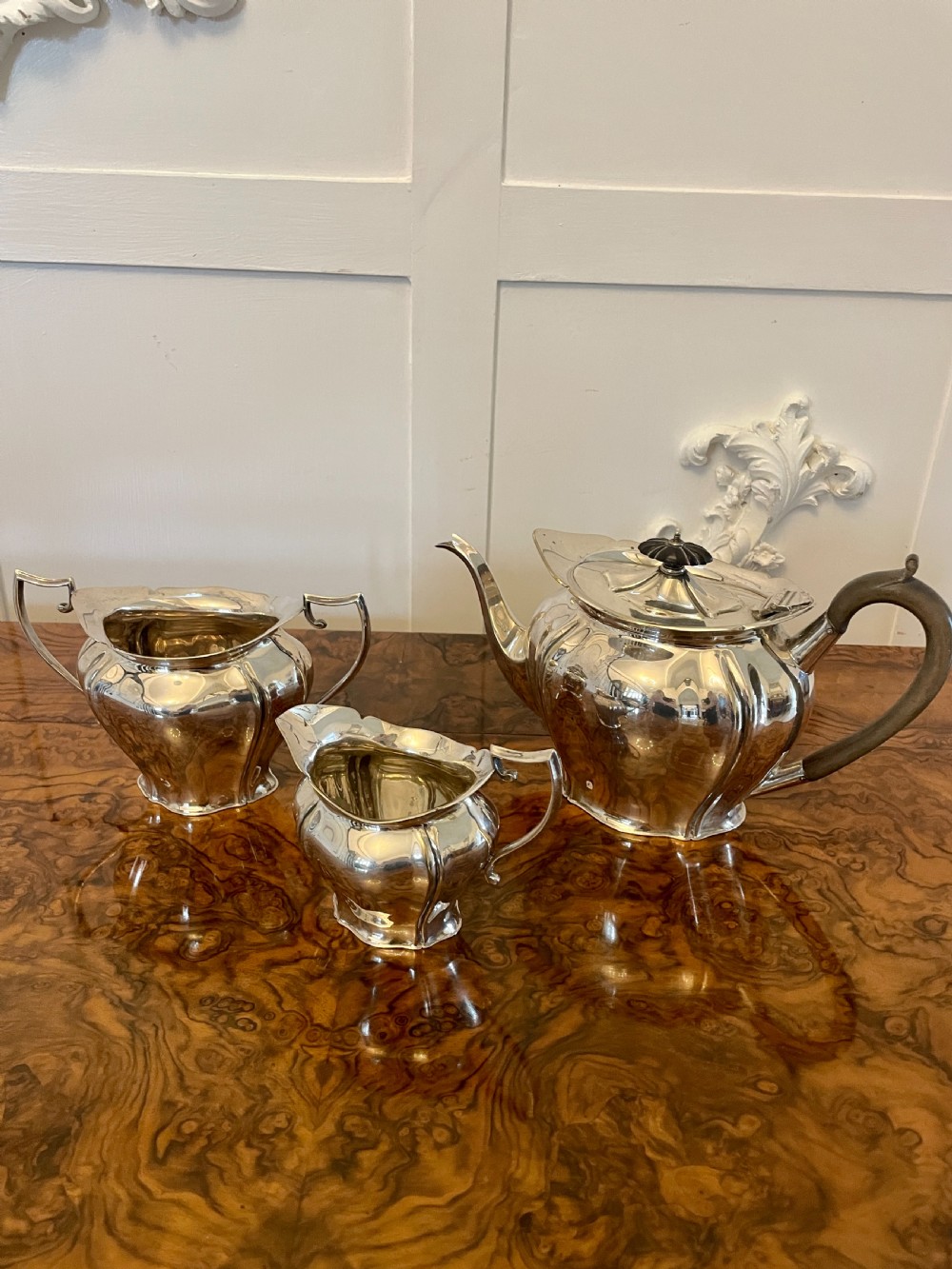 A three-piece Edwardian silver-plated tea set featuring a fluted teapot with a dark wooden handle, a matching double-handled sugar bowl, and a cream jug.