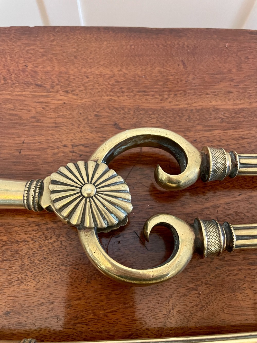 A set of three antique brass fire irons including tongs, a poker, and an engraved shovel, laid flat on a dark wooden surface. View II