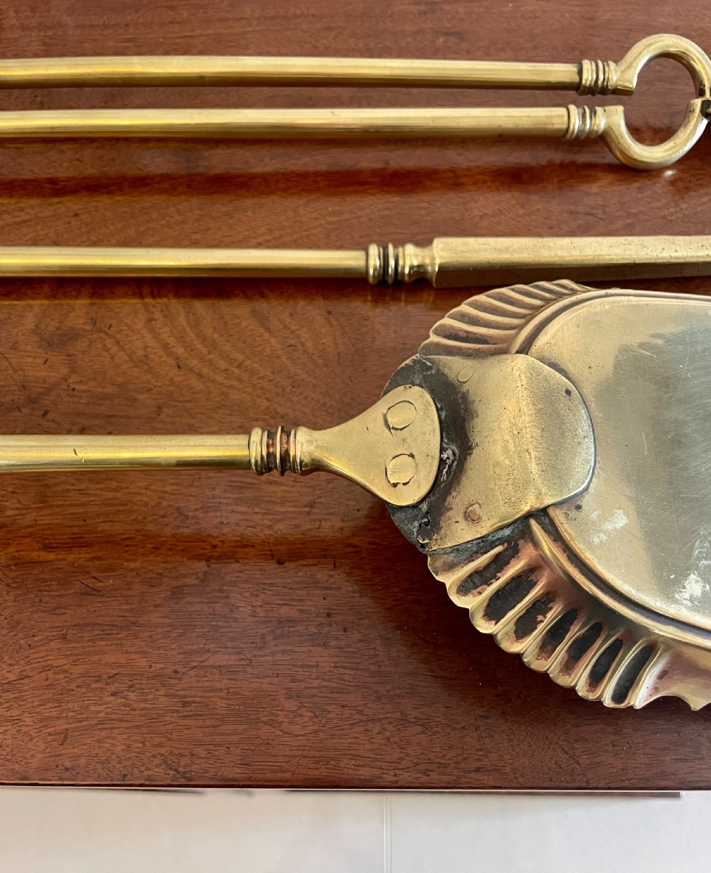 A set of three antique brass fire irons including tongs, a poker, and an engraved shovel, laid flat on a dark wooden surface. View III