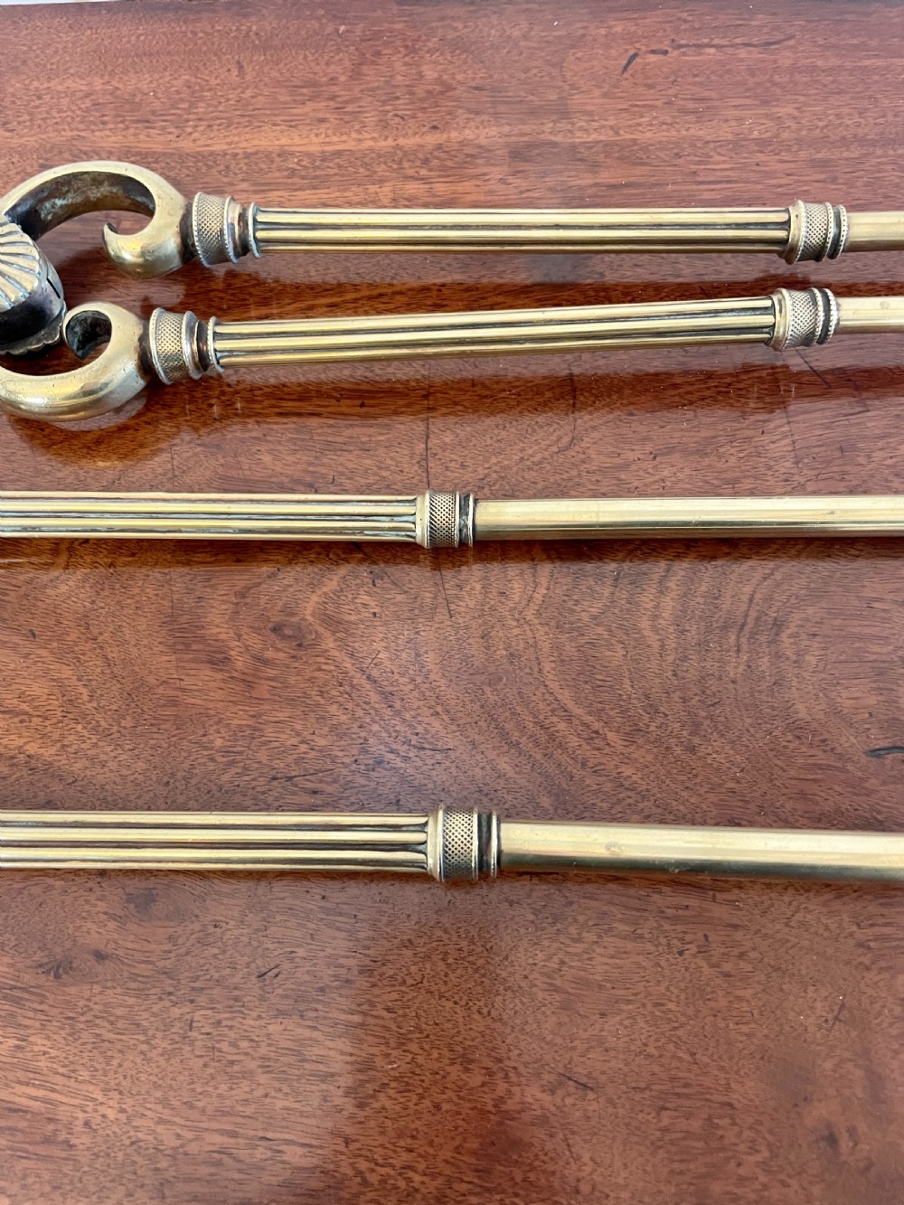 A set of three antique brass fire irons including tongs, a poker, and an engraved shovel, laid flat on a dark wooden surface. View V