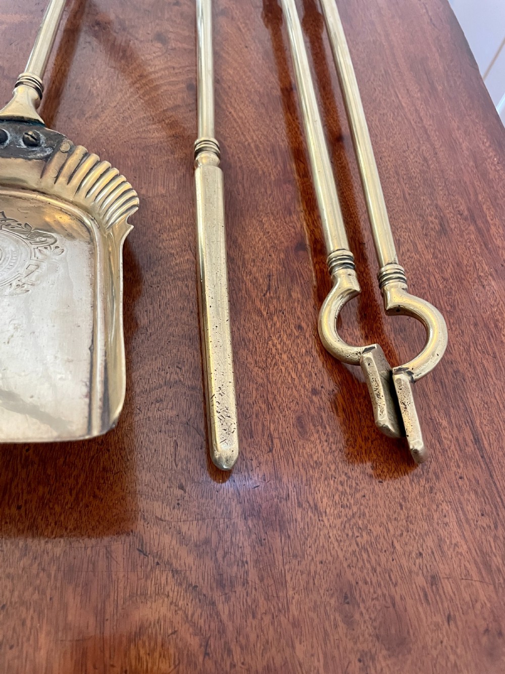 A set of three antique brass fire irons including tongs, a poker, and an engraved shovel, laid flat on a dark wooden surface. View VI