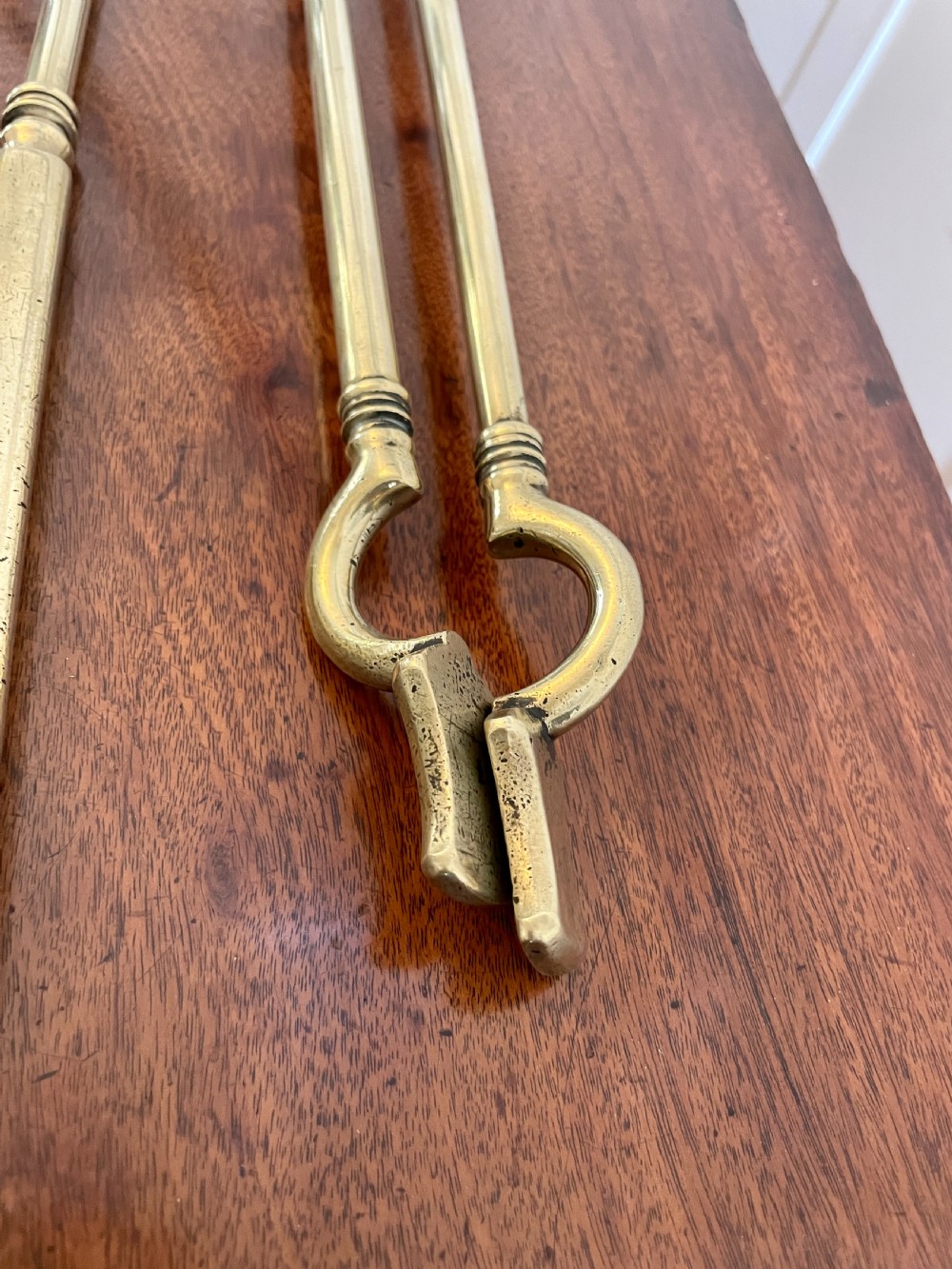 A set of three antique brass fire irons including tongs, a poker, and an engraved shovel, laid flat on a dark wooden surface. View VII