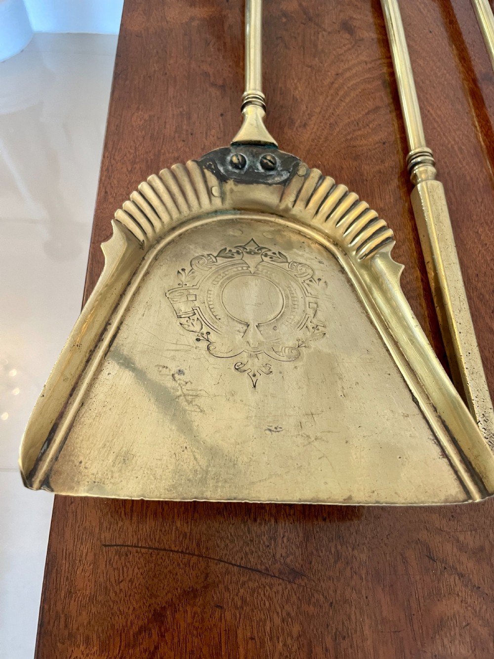 A set of three antique brass fire irons including tongs, a poker, and an engraved shovel, laid flat on a dark wooden surface. View VIII