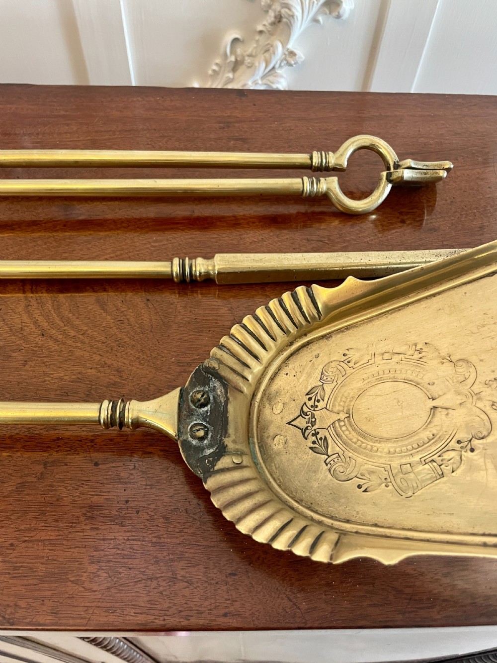 A set of three antique brass fire irons including tongs, a poker, and an engraved shovel, laid flat on a dark wooden surface. View IX