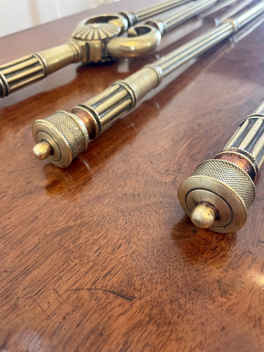 A set of three antique brass fire irons including tongs, a poker, and an engraved shovel, laid flat on a dark wooden surface. View X