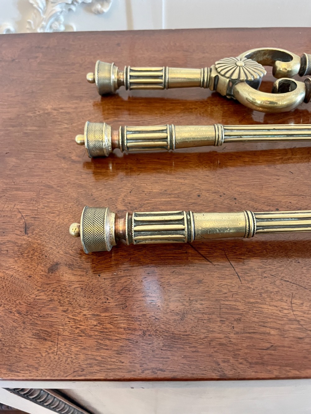 A set of three antique brass fire irons including tongs, a poker, and an engraved shovel, laid flat on a dark wooden surface. View XI