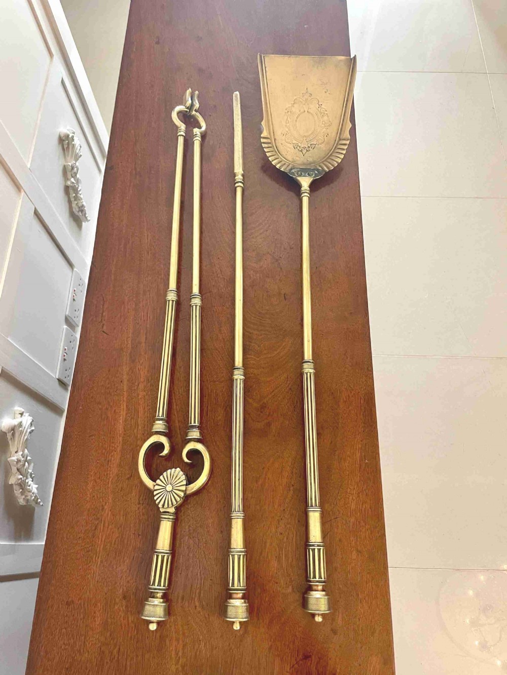 A set of three antique brass fire irons including tongs, a poker, and an engraved shovel, laid flat on a dark wooden surface.