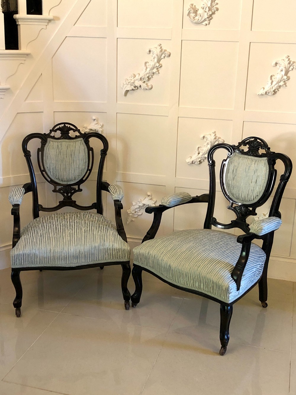 A pair of Victorian carved black lacquered open armchairs featuring oval padded backrests, curved padded arms, pale green striped fabric, and cabriole legs with brass castors. View II