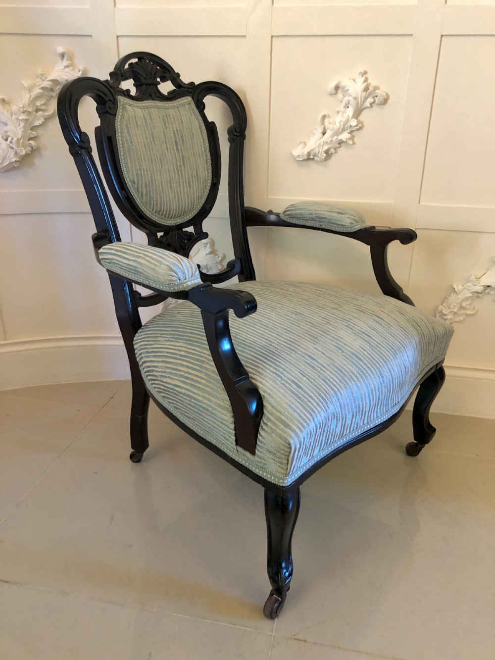A pair of Victorian carved black lacquered open armchairs featuring oval padded backrests, curved padded arms, pale green striped fabric, and cabriole legs with brass castors. View XIII