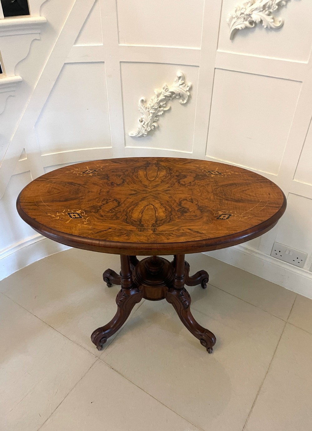 An antique Victorian oval centre table featuring book-matched burr walnut veneer with intricate floral inlay, supported by a carved central pedestal and four splayed legs on castors.