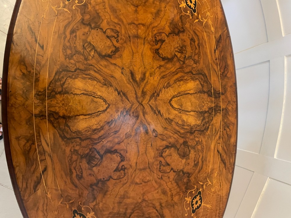 An antique Victorian oval centre table featuring book-matched burr walnut veneer with intricate floral inlay, supported by a carved central pedestal and four splayed legs on castors. View VIII