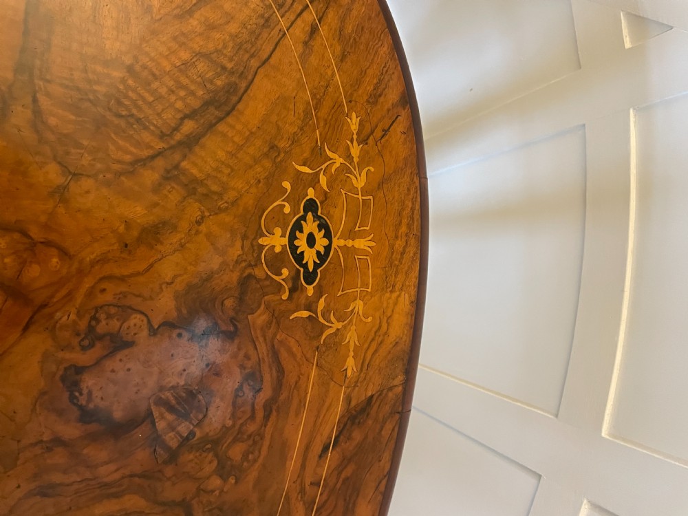 An antique Victorian oval centre table featuring book-matched burr walnut veneer with intricate floral inlay, supported by a carved central pedestal and four splayed legs on castors. View IX