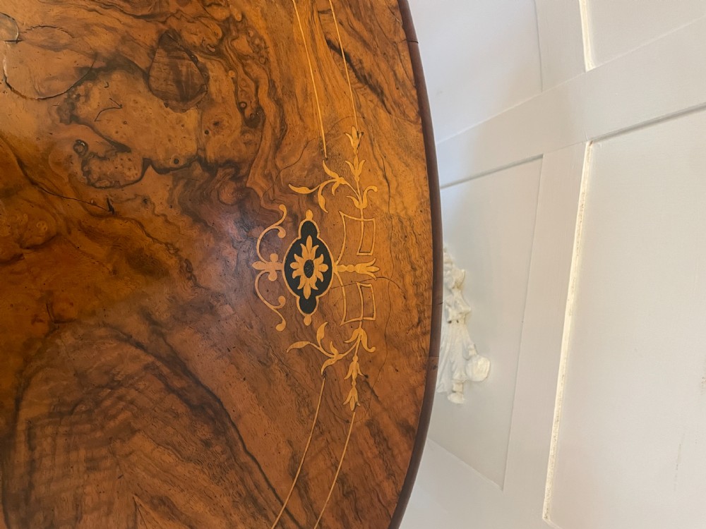 An antique Victorian oval centre table featuring book-matched burr walnut veneer with intricate floral inlay, supported by a carved central pedestal and four splayed legs on castors. View X