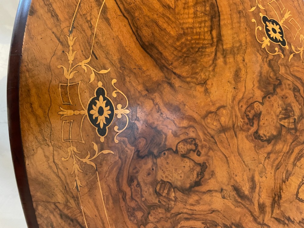 An antique Victorian oval centre table featuring book-matched burr walnut veneer with intricate floral inlay, supported by a carved central pedestal and four splayed legs on castors. View XII