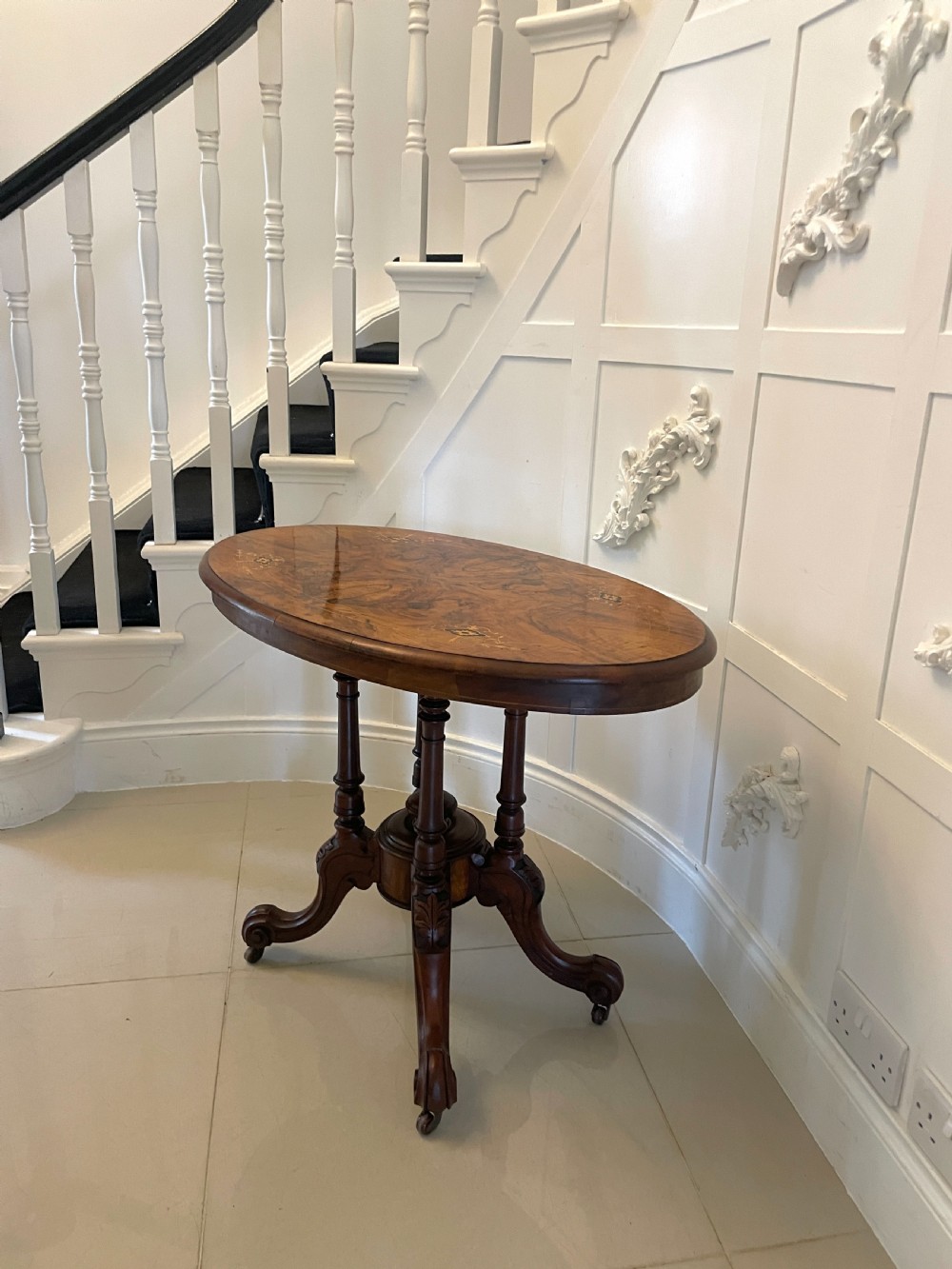 An antique Victorian oval centre table featuring book-matched burr walnut veneer with intricate floral inlay, supported by a carved central pedestal and four splayed legs on castors. View XIV