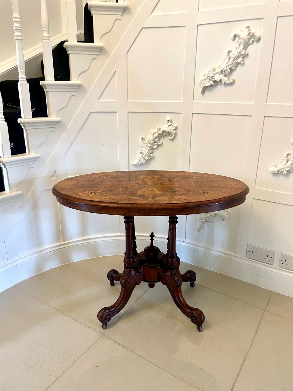 An antique Victorian oval centre table featuring book-matched burr walnut veneer with intricate floral inlay, supported by a carved central pedestal and four splayed legs on castors. View XV