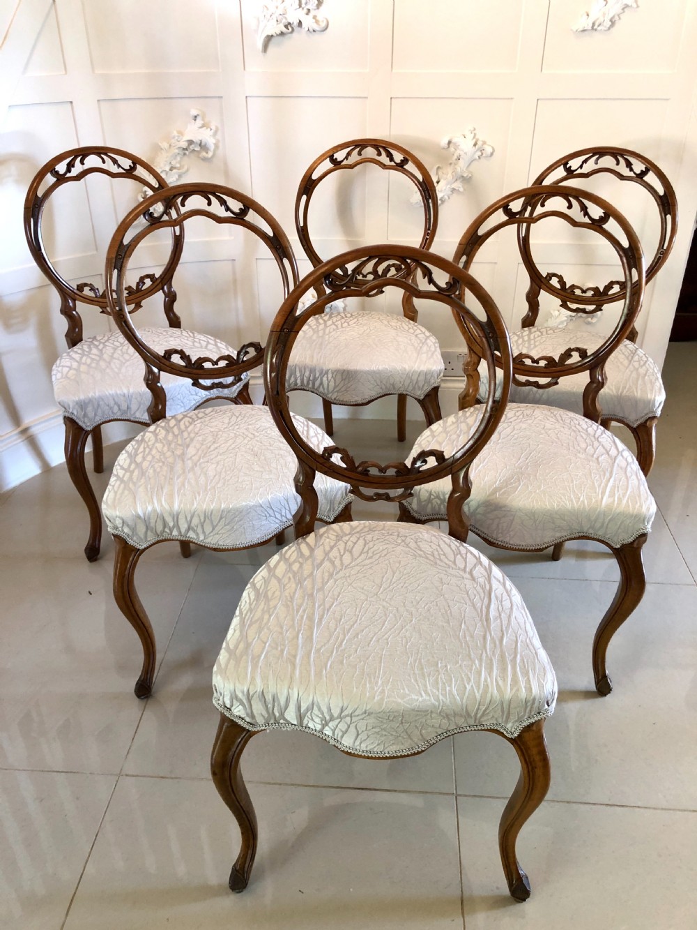 A set of six Victorian walnut dining chairs featuring carved circular balloon backs, cabriole front legs, and light silver patterned upholstered seats.