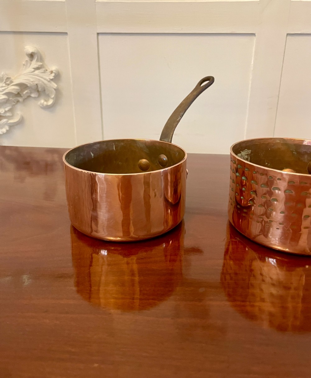 A set of three graduated Victorian copper saucepans featuring dark iron handles resting on a polished wooden surface. View X