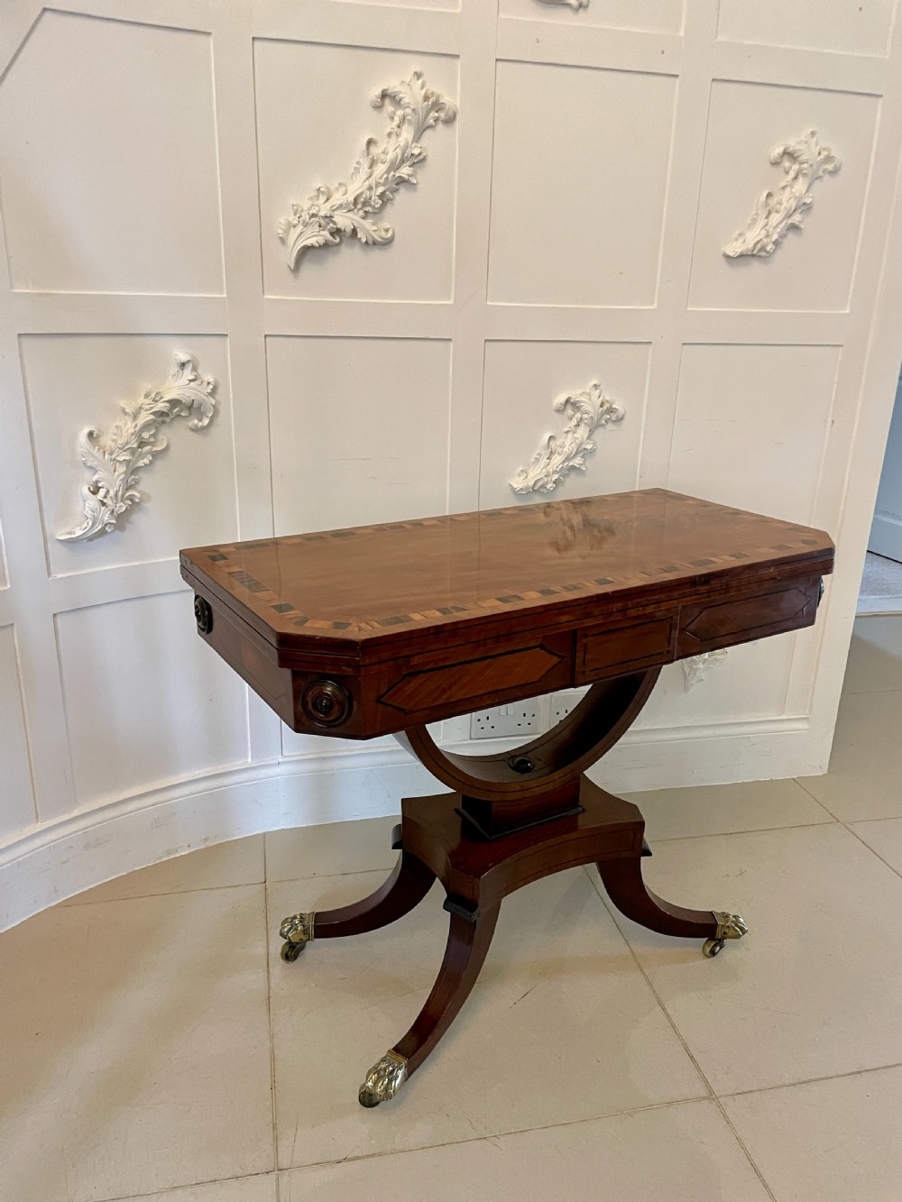 A Regency mahogany fold-over card table featuring crossbanded inlay, a central pedestal base, and four splayed legs with brass paw castors. View V