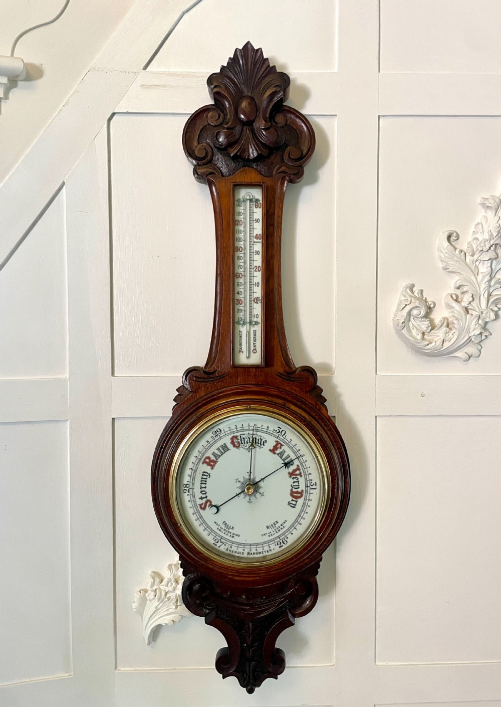 An upright Victorian carved oak banjo barometer featuring a central thermometer column and a circular aneroid dial at the base.