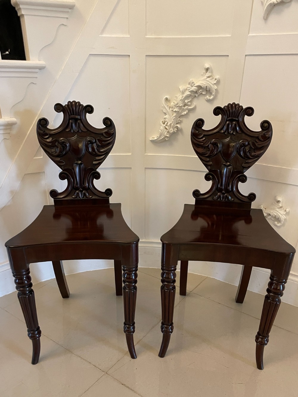 A pair of dark mahogany Regency hall chairs featuring intricate carved cartouche backs, shaped solid wooden seats, and turned, reeded front legs.