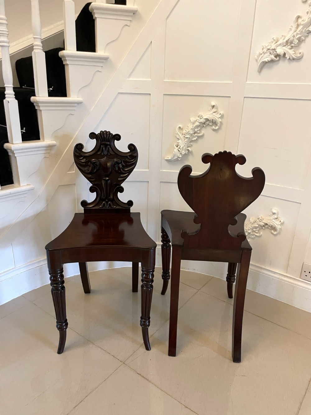 A pair of dark mahogany Regency hall chairs featuring intricate carved cartouche backs, shaped solid wooden seats, and turned, reeded front legs. View VII