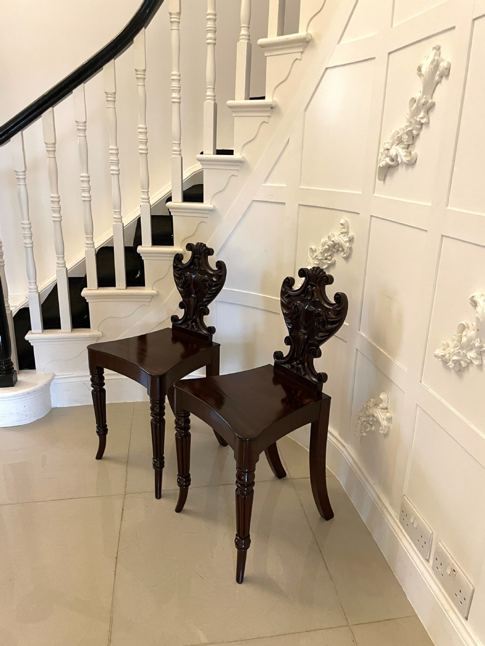 A pair of dark mahogany Regency hall chairs featuring intricate carved cartouche backs, shaped solid wooden seats, and turned, reeded front legs. View VIII