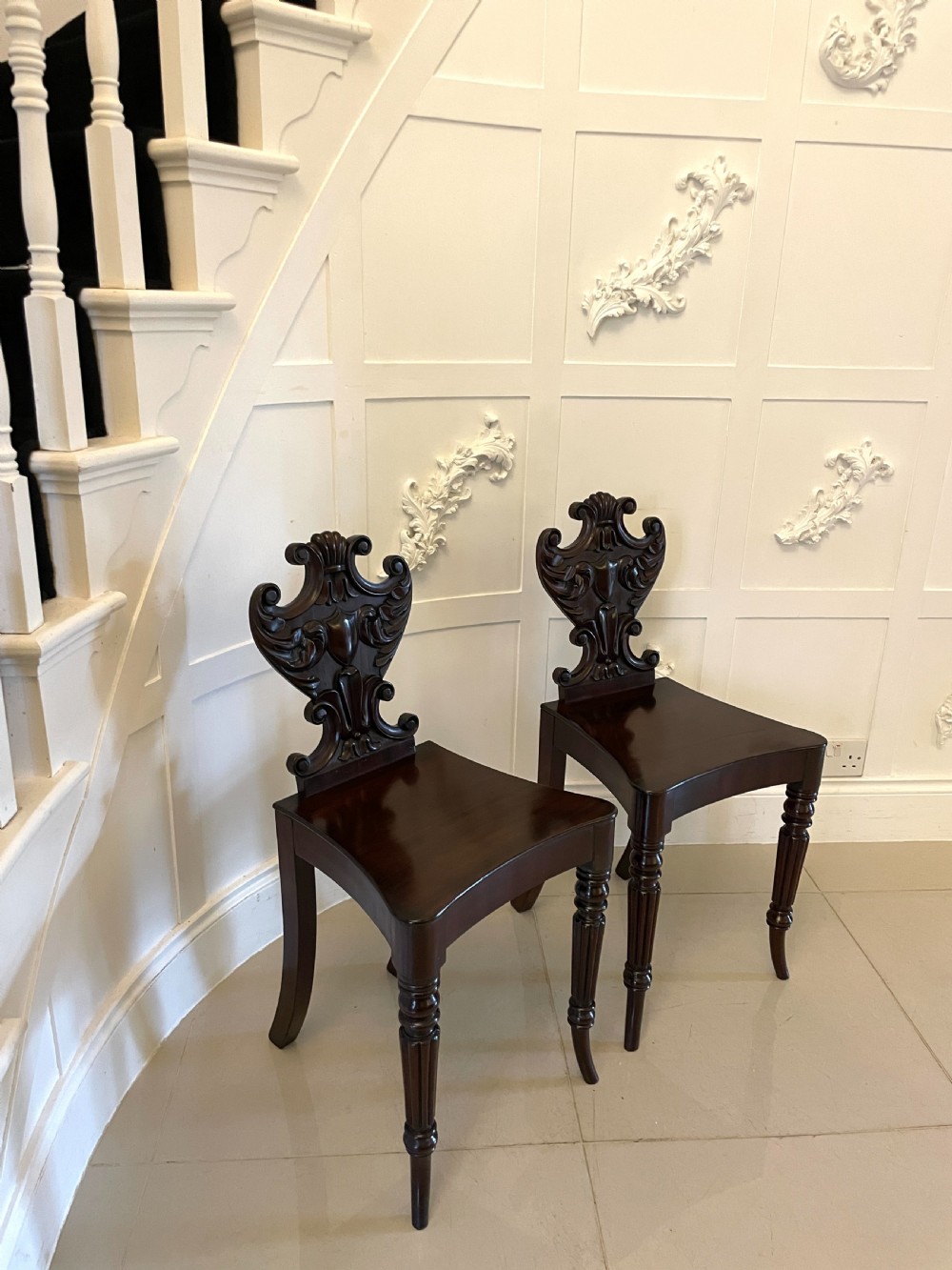 A pair of dark mahogany Regency hall chairs featuring intricate carved cartouche backs, shaped solid wooden seats, and turned, reeded front legs. View IX