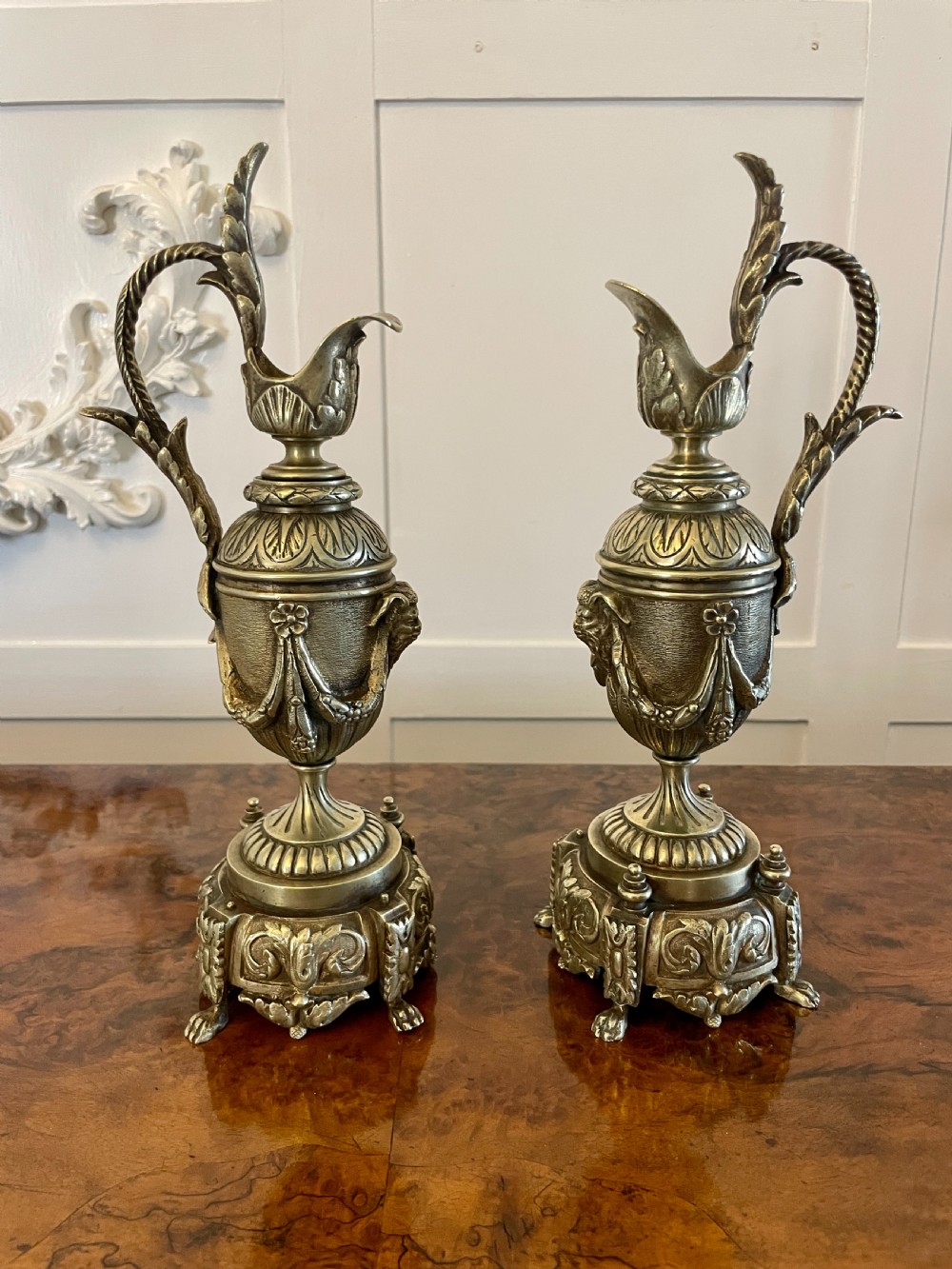 A matching pair of ornate Victorian brass ewers with decorative scroll handles, leaf motifs, and footed bases resting on a polished timber surface.
