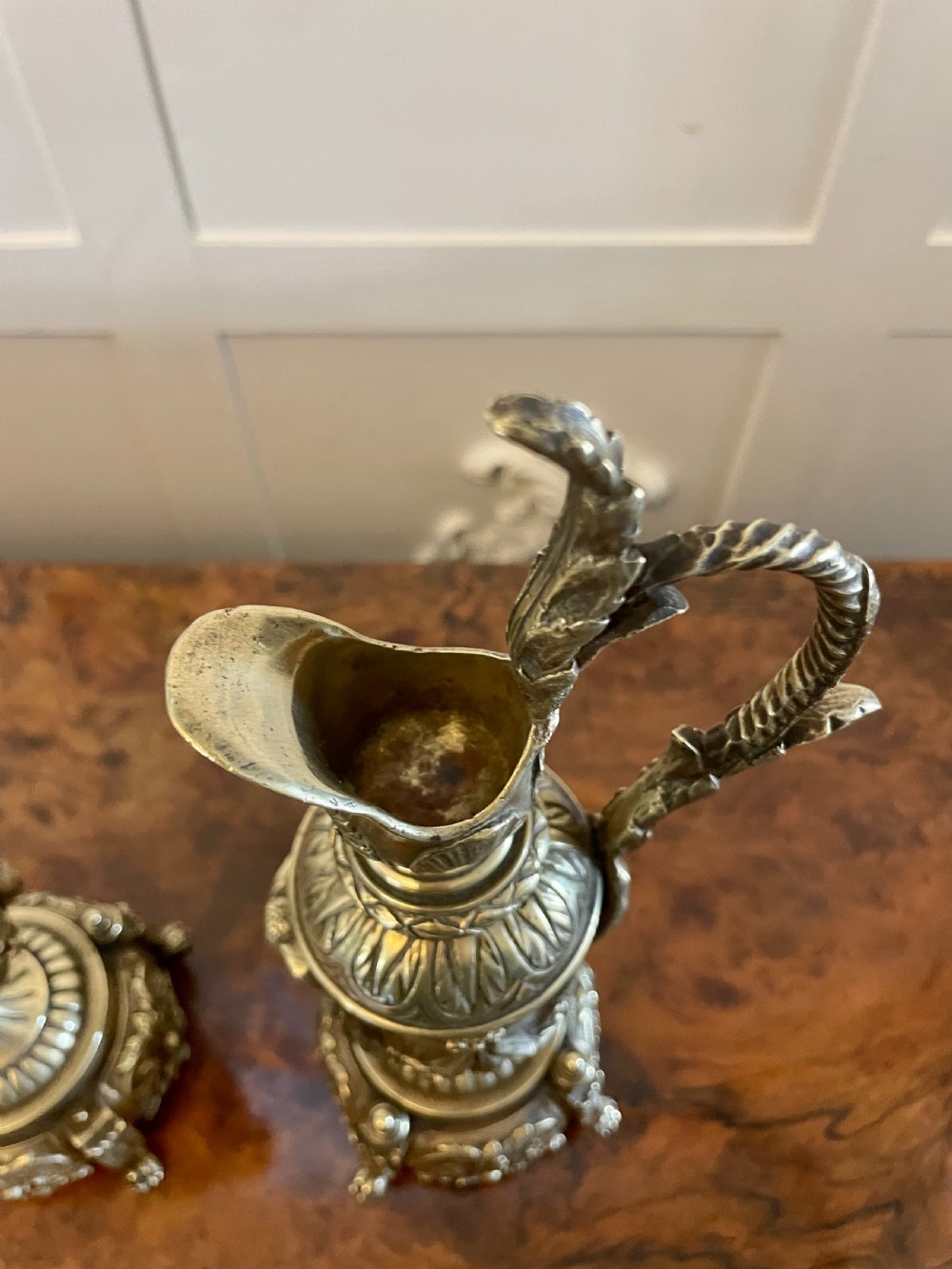 A matching pair of ornate Victorian brass ewers with decorative scroll handles, leaf motifs, and footed bases resting on a polished timber surface. View III