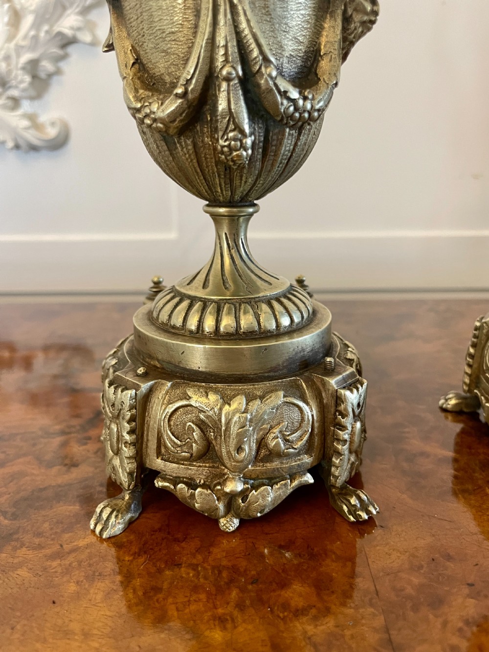A matching pair of ornate Victorian brass ewers with decorative scroll handles, leaf motifs, and footed bases resting on a polished timber surface. View V