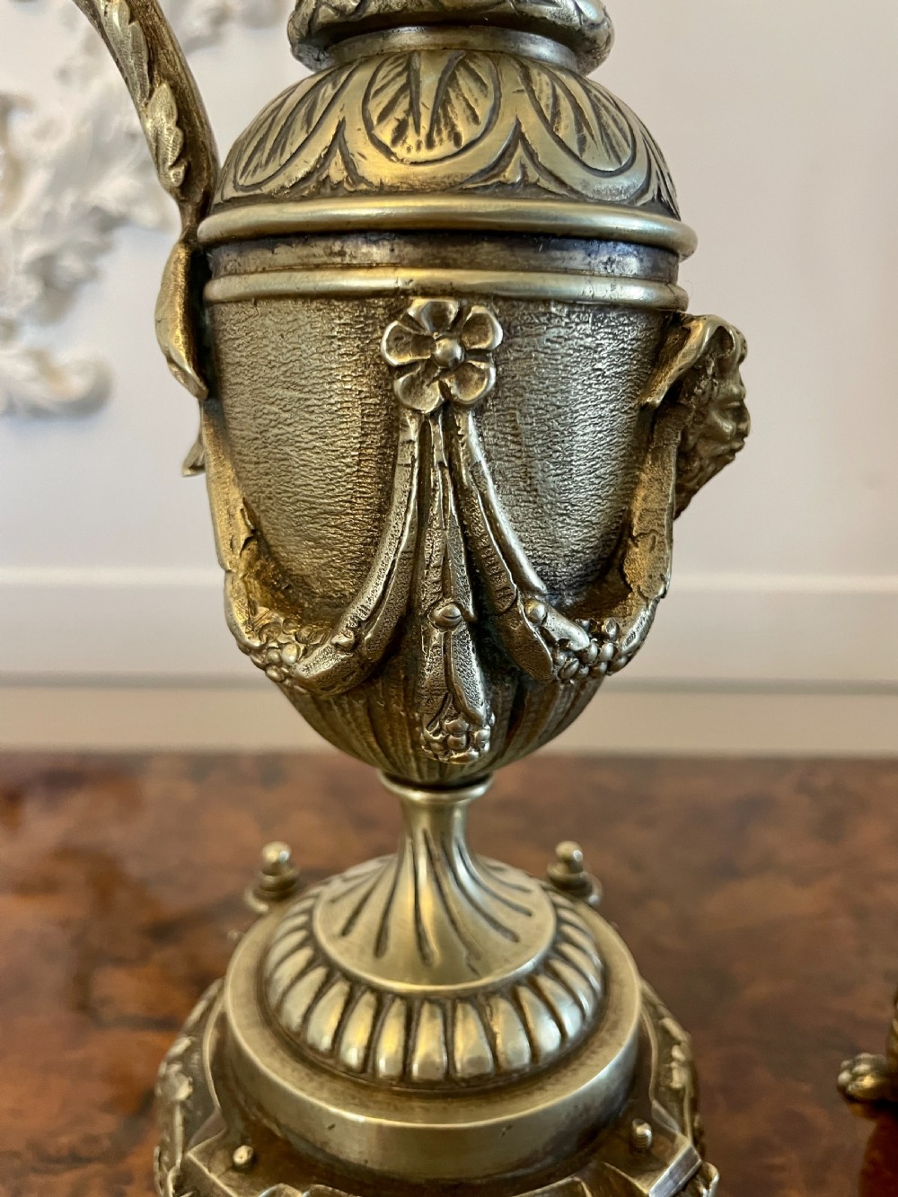 A matching pair of ornate Victorian brass ewers with decorative scroll handles, leaf motifs, and footed bases resting on a polished timber surface. View VI
