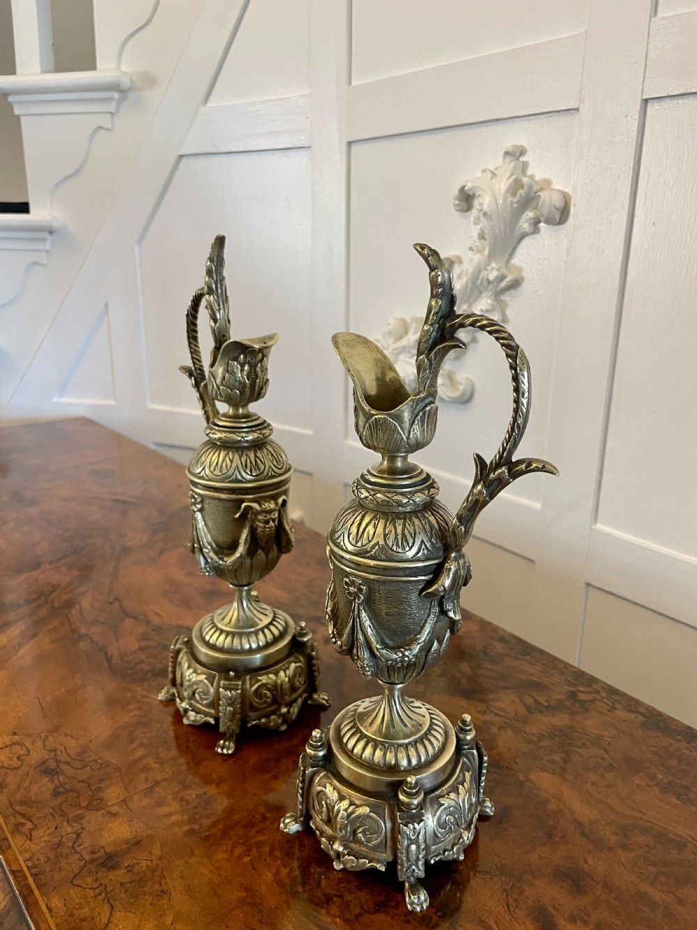 A matching pair of ornate Victorian brass ewers with decorative scroll handles, leaf motifs, and footed bases resting on a polished timber surface. View VIII