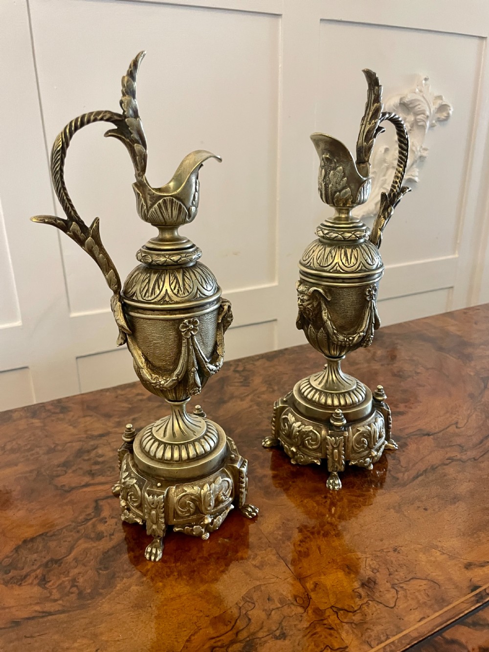 A matching pair of ornate Victorian brass ewers with decorative scroll handles, leaf motifs, and footed bases resting on a polished timber surface. View IX