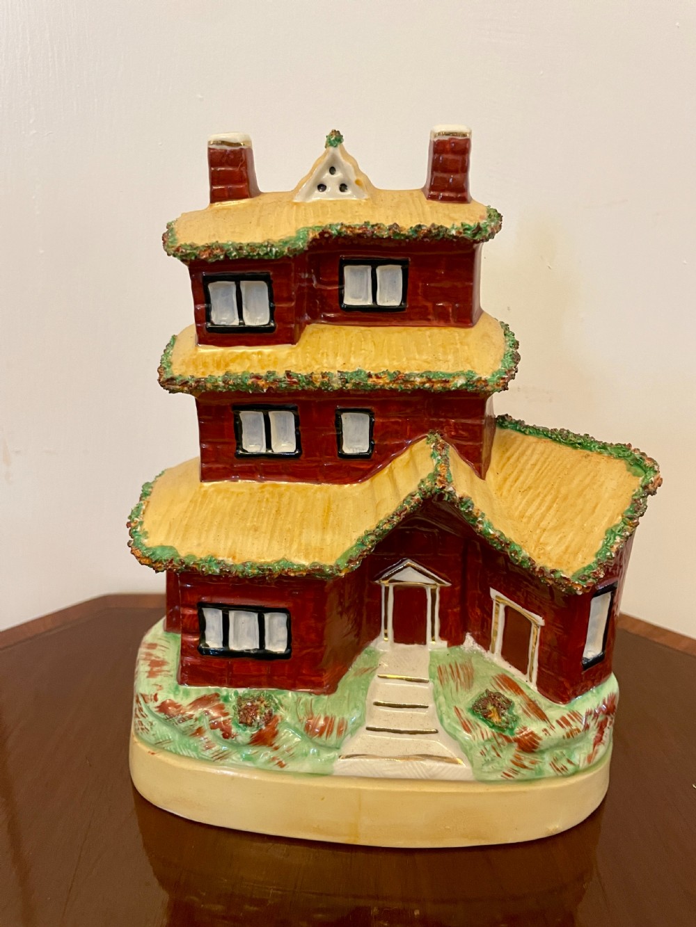 A Victorian Staffordshire pottery flatback figurine depicting a multi-storey terracotta red house with a yellow thatched roof, green encrusted foliage trim, and two chimneys.