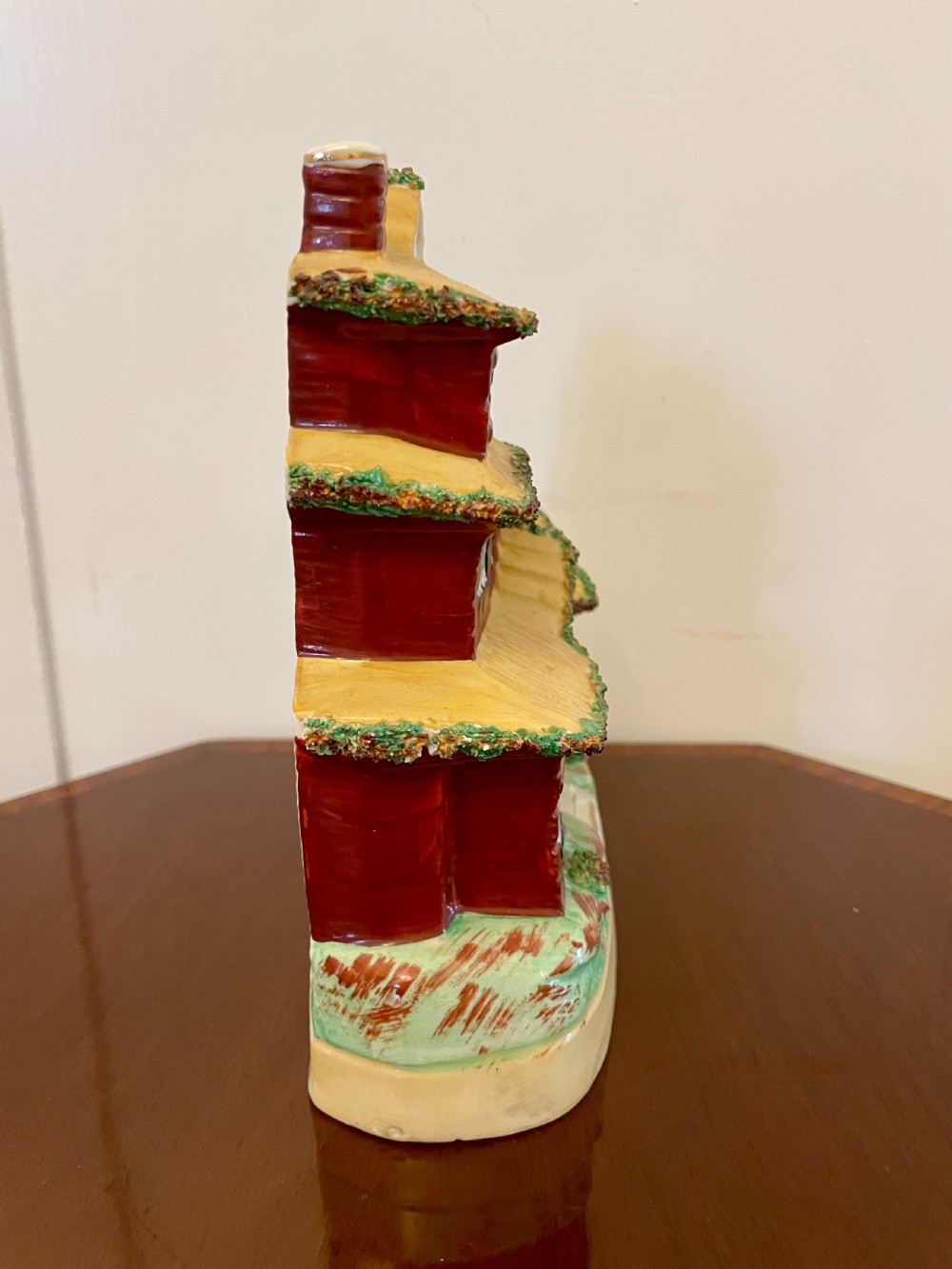 A Victorian Staffordshire pottery flatback figurine depicting a multi-storey terracotta red house with a yellow thatched roof, green encrusted foliage trim, and two chimneys. View VIII
