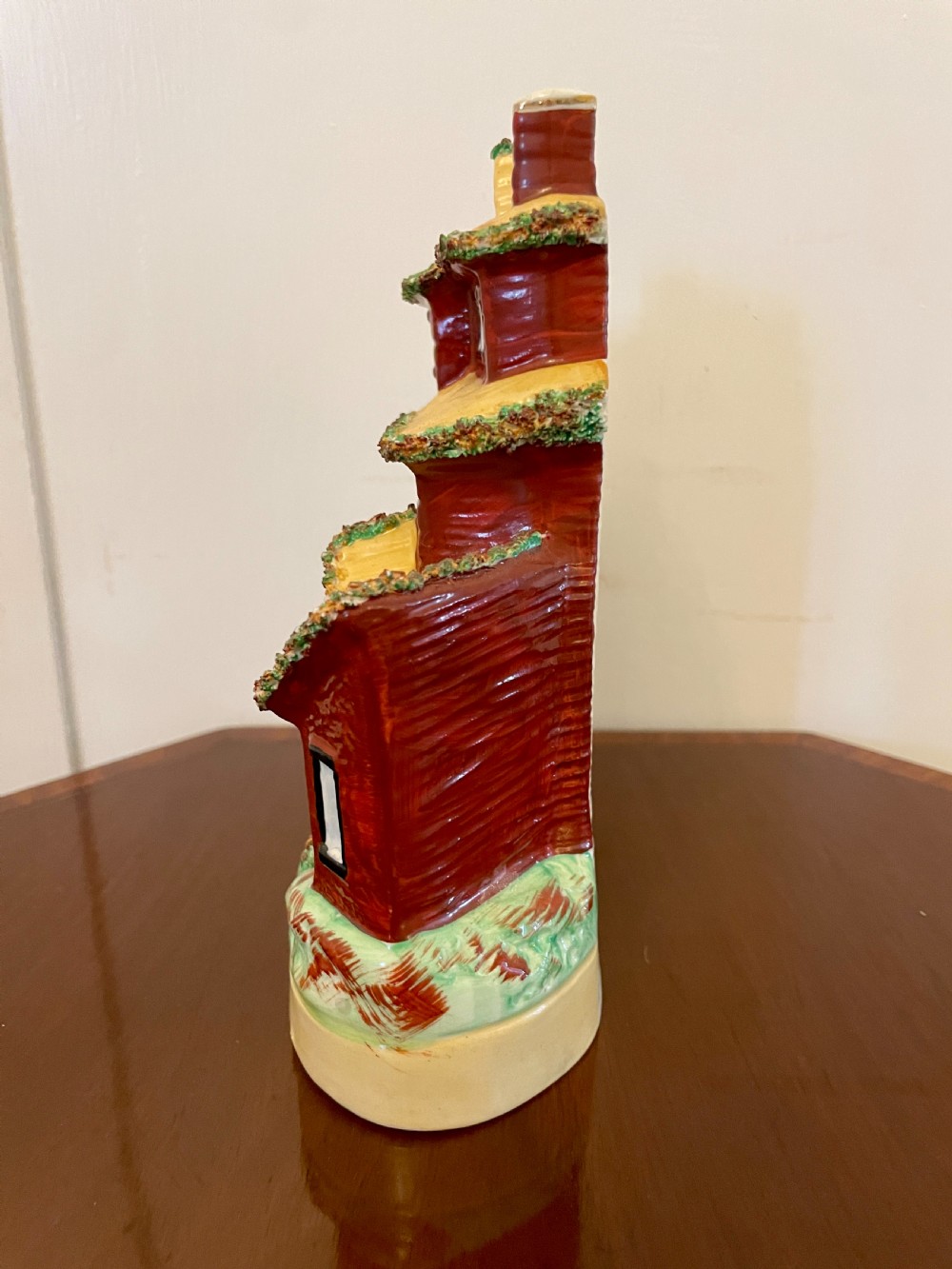 A Victorian Staffordshire pottery flatback figurine depicting a multi-storey terracotta red house with a yellow thatched roof, green encrusted foliage trim, and two chimneys. View IX