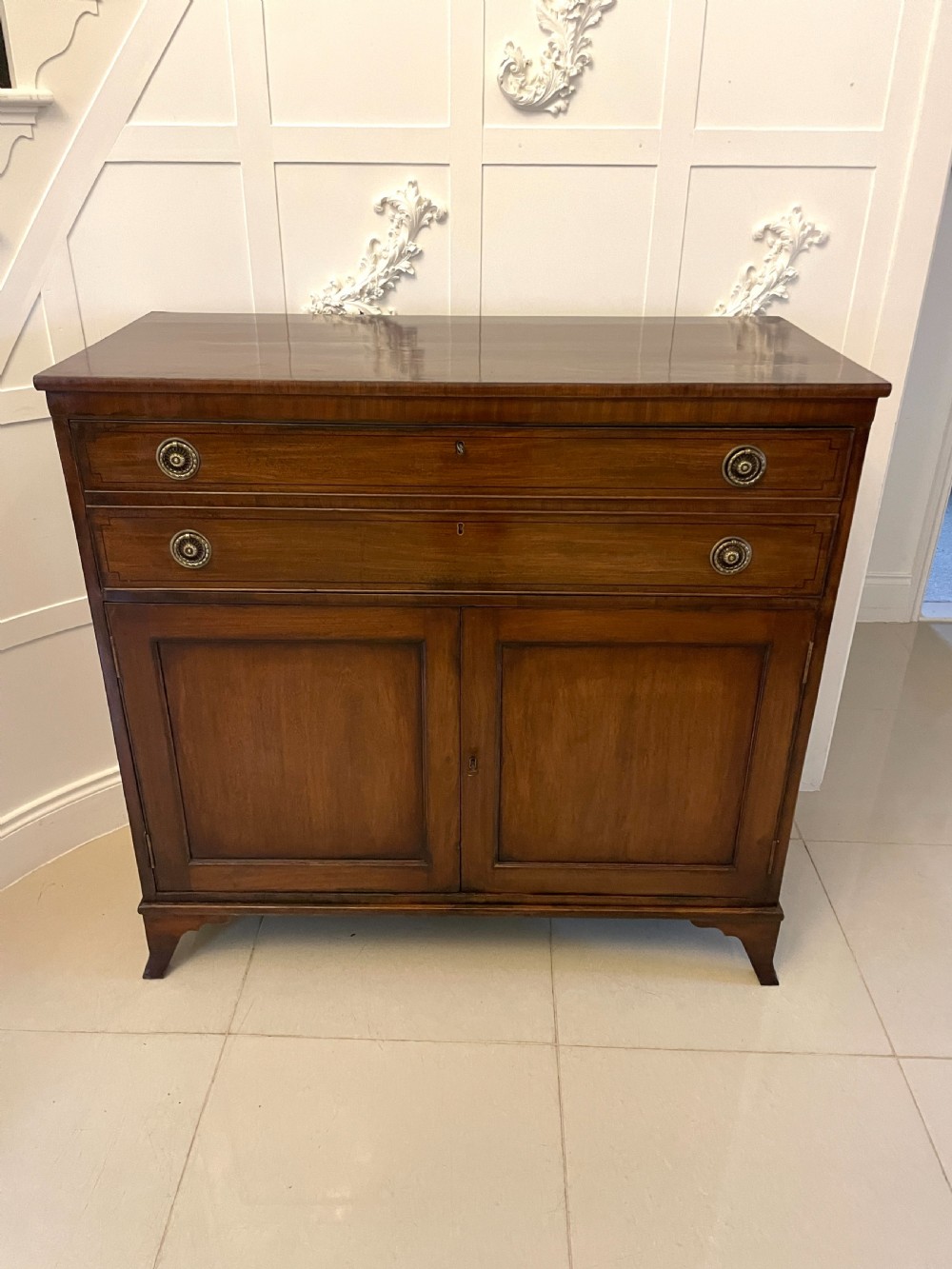 A George III mahogany secretaire desk featuring top drawers with brass ring handles over a two-door panelled cupboard base resting on bracket feet.