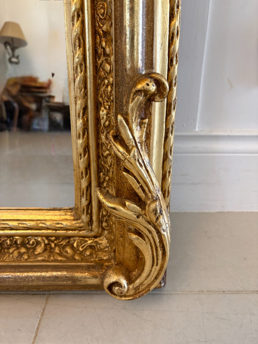 A large rectangular wall mirror featuring an ornate gilded frame decorated with carved floral scrolls and two cherub figures atop the crest. View IV
