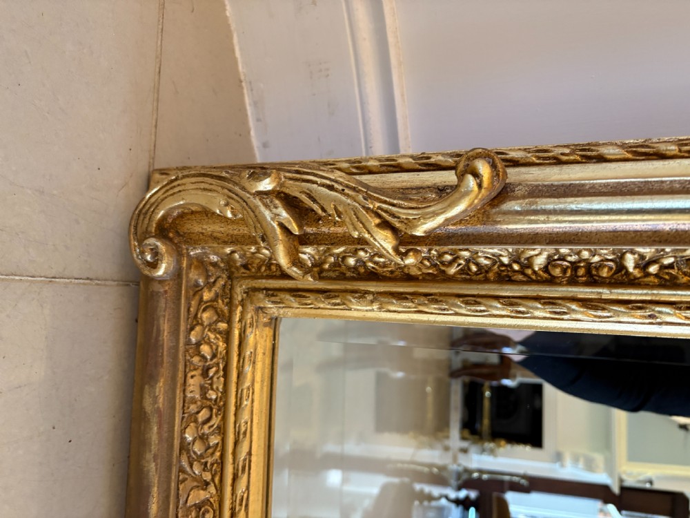 A large rectangular wall mirror featuring an ornate gilded frame decorated with carved floral scrolls and two cherub figures atop the crest. View V