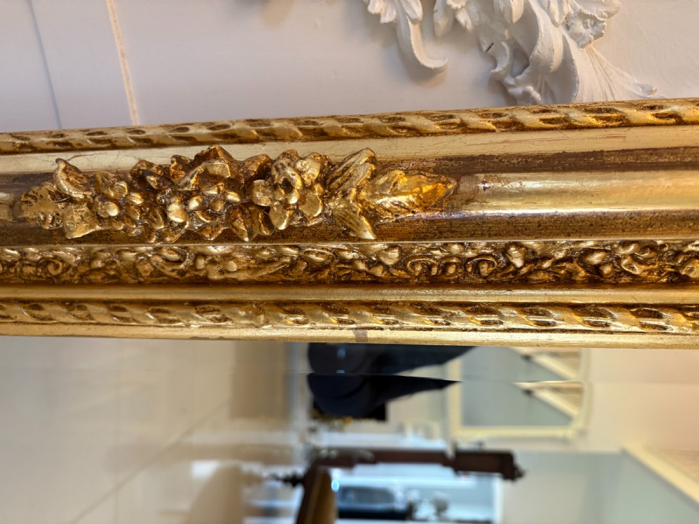 A large rectangular wall mirror featuring an ornate gilded frame decorated with carved floral scrolls and two cherub figures atop the crest. View VI