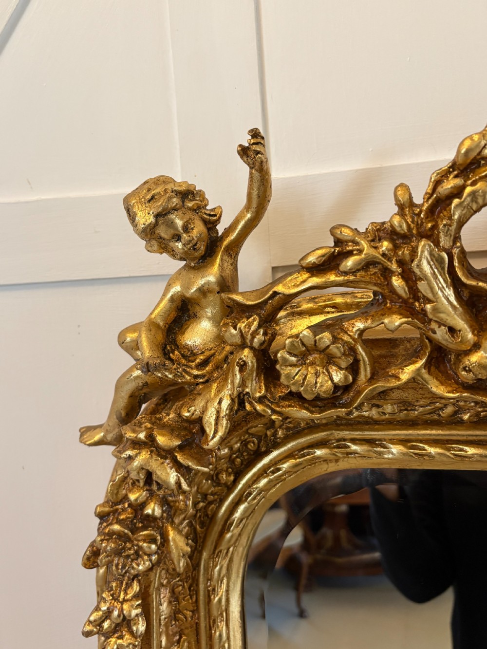 A large rectangular wall mirror featuring an ornate gilded frame decorated with carved floral scrolls and two cherub figures atop the crest. View VII