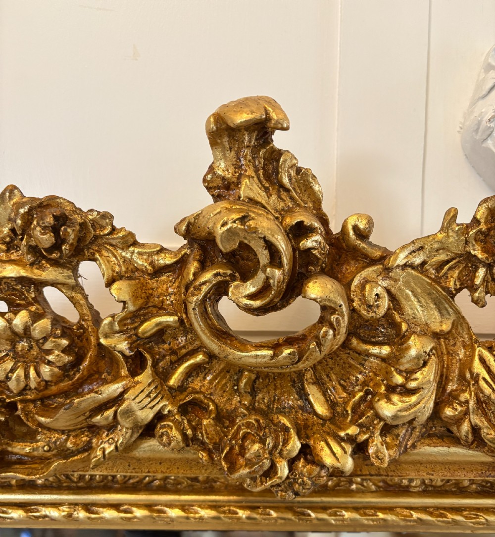A large rectangular wall mirror featuring an ornate gilded frame decorated with carved floral scrolls and two cherub figures atop the crest. View VIII