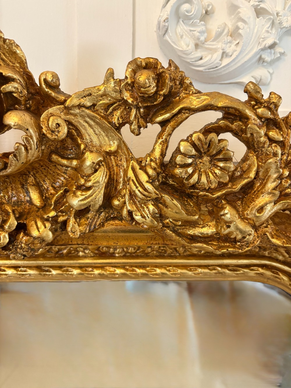 A large rectangular wall mirror featuring an ornate gilded frame decorated with carved floral scrolls and two cherub figures atop the crest. View IX