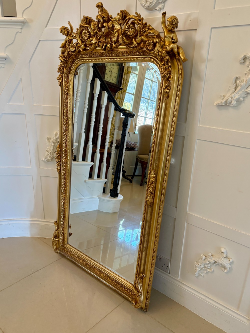 A large rectangular wall mirror featuring an ornate gilded frame decorated with carved floral scrolls and two cherub figures atop the crest. View XI