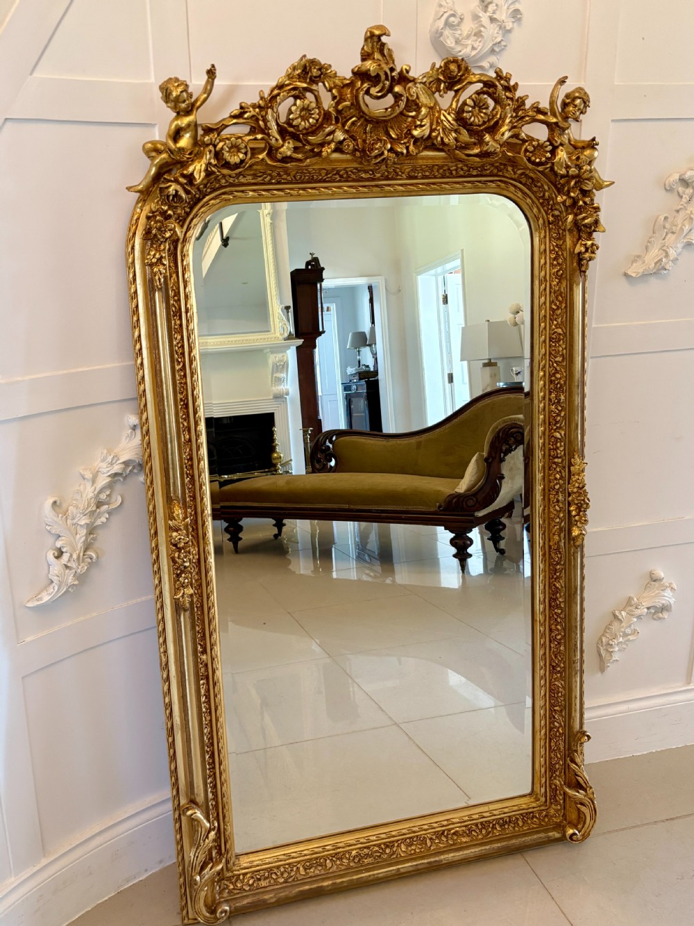 A large rectangular wall mirror featuring an ornate gilded frame decorated with carved floral scrolls and two cherub figures atop the crest. View XII