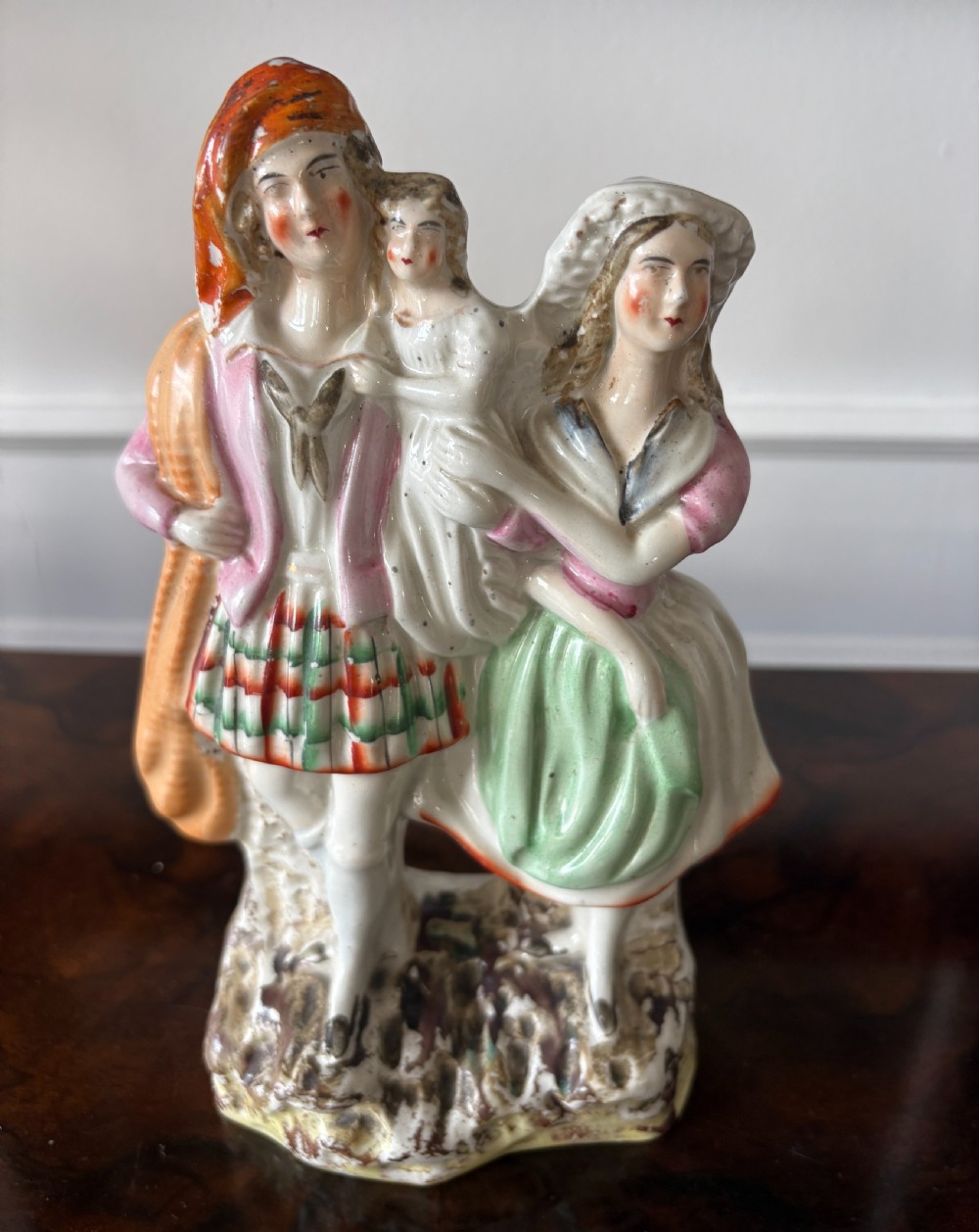 Victorian Staffordshire earthenware figurine depicting a man in a tartan kilt holding a child next to a woman, painted in polychrome glazes on a moulded base.