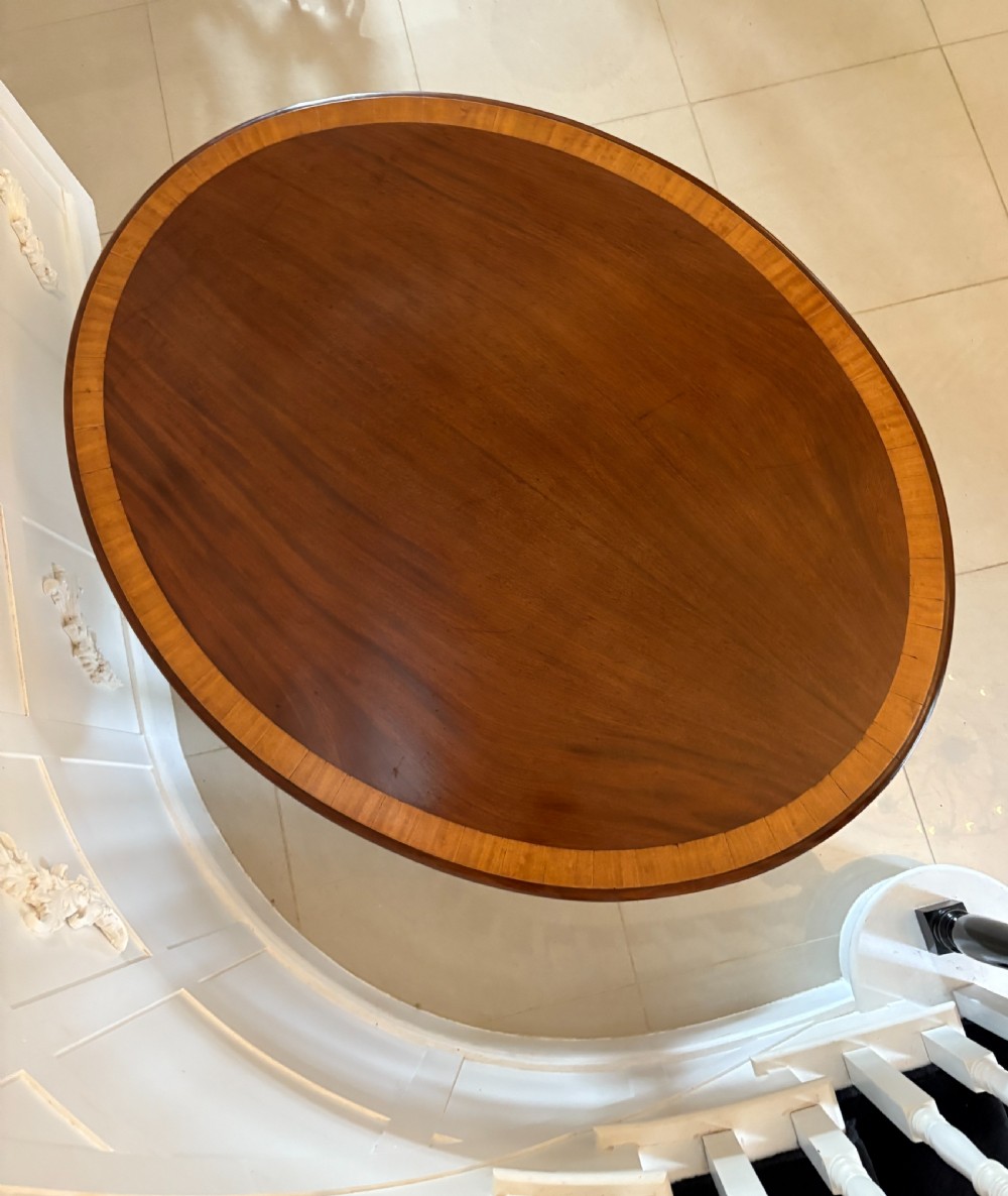 An antique Victorian oval mahogany dining table featuring crossbanded satinwood inlay, shown tilted vertically on a carved quadruped base with scroll feet and brass castors. View II